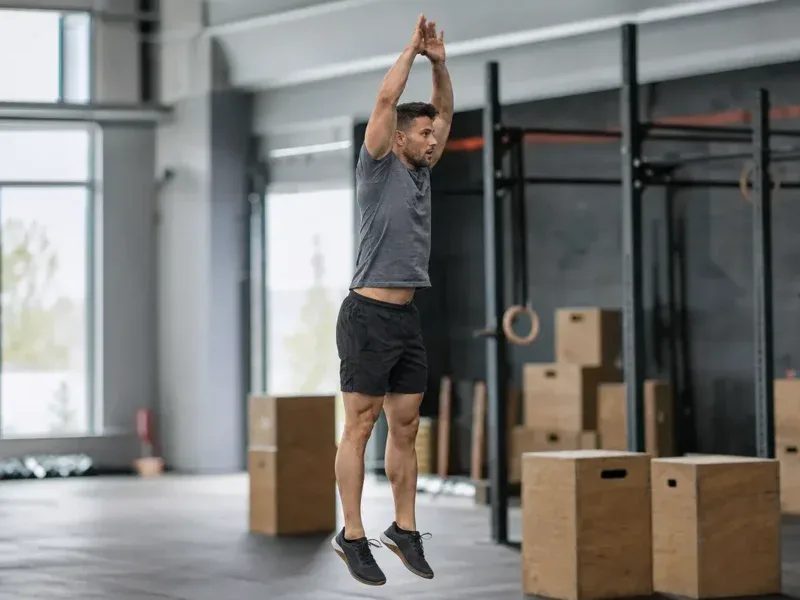 Mężczyzna w wyskoku z klaśnięciem nad głową kończy powtórzenie burpee w boksie crossfitowym