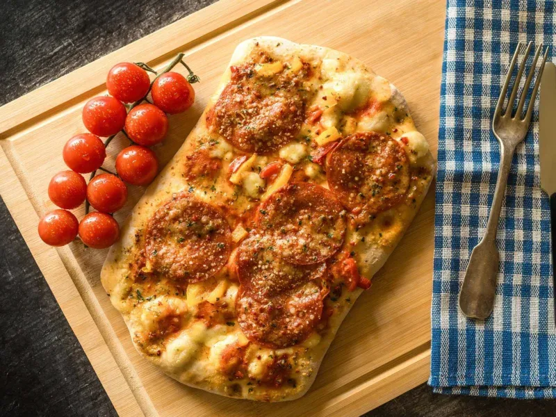 Pizza z salami na drewnianej desce, obok pomidorki koktajlowe i sztućce