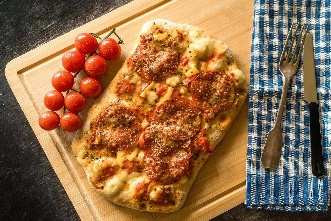Pizza z salami na drewnianej desce, obok pomidorki koktajlowe i sztućce